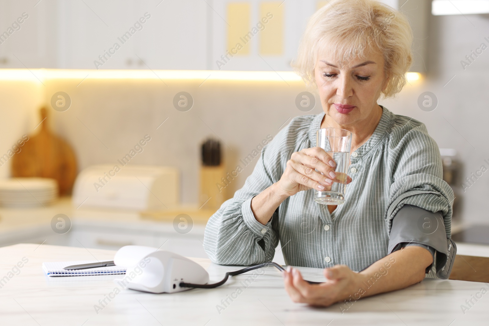 Photo of Senior woman with glass of water measuring blood pressure at table in kitchen. Space for text