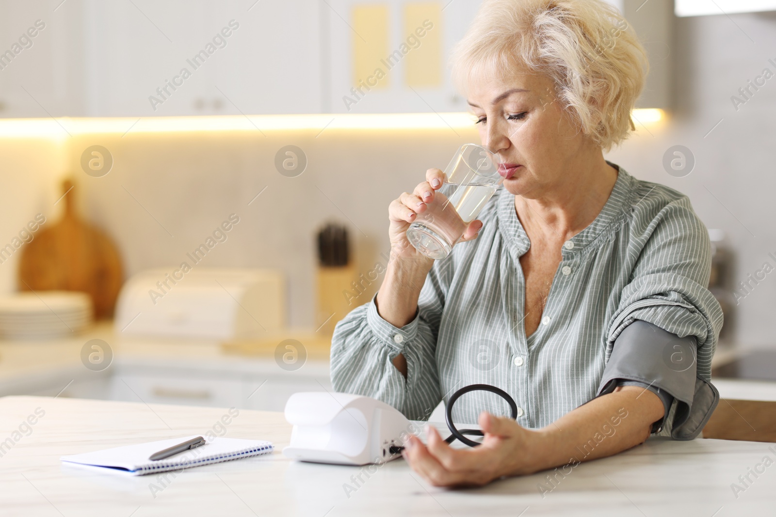 Photo of Senior woman with glass of water measuring blood pressure at table in kitchen. Space for text