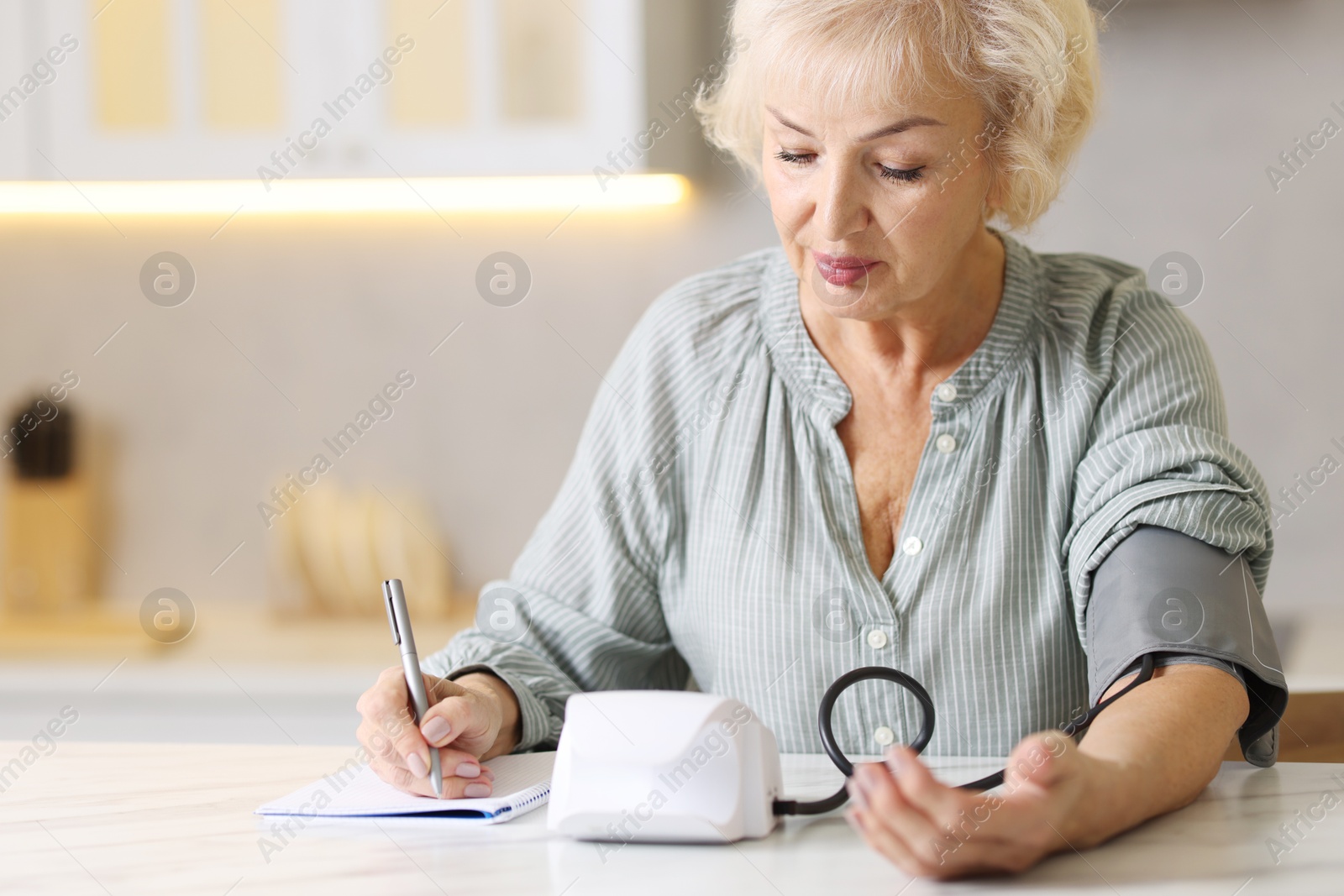 Photo of Senior woman writing results of blood pressure measurement in kitchen at home. Space for text