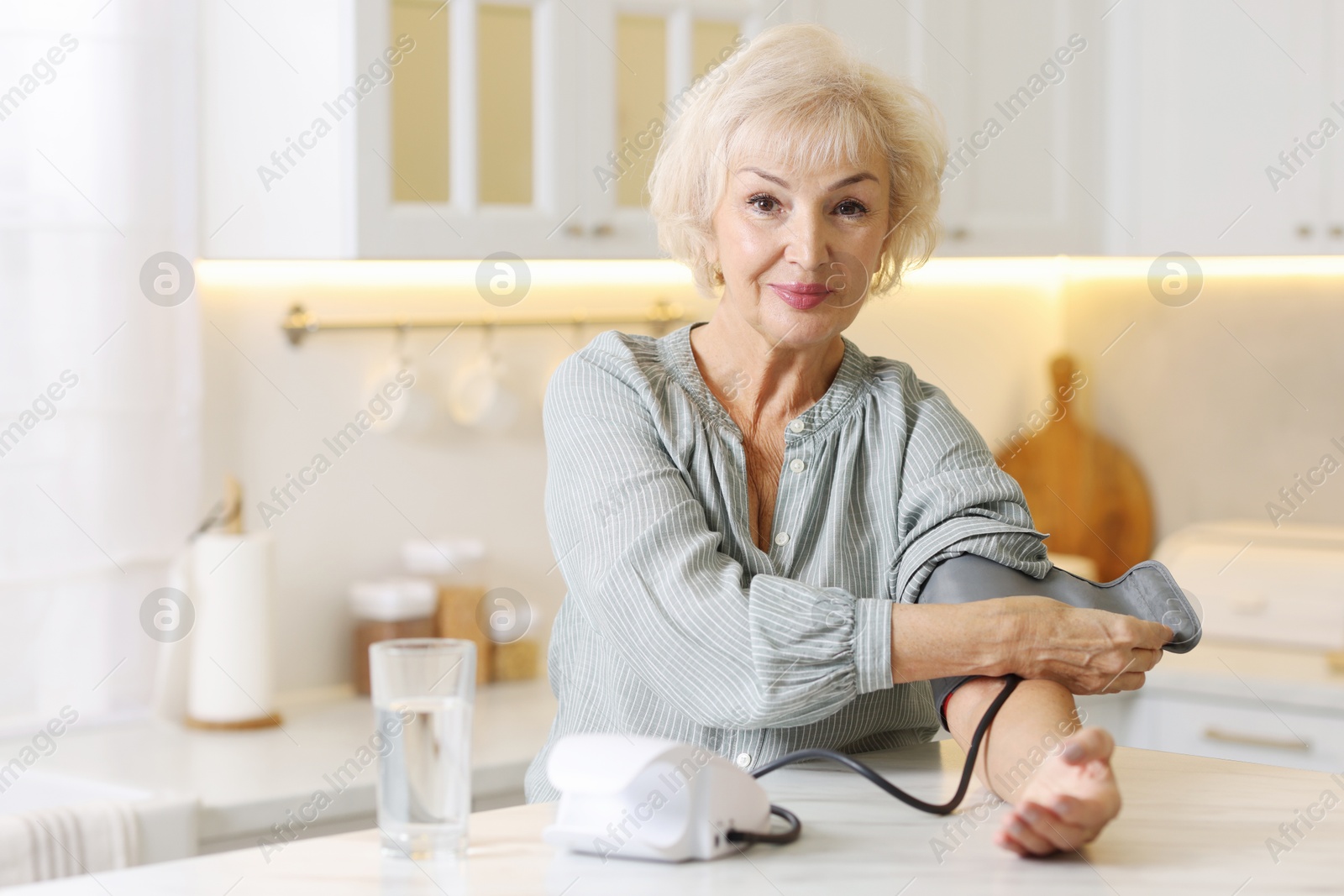 Photo of Senior woman measuring blood pressure at table in kitchen