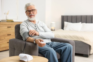 Senior man measuring blood pressure in armchair at home. Space for text