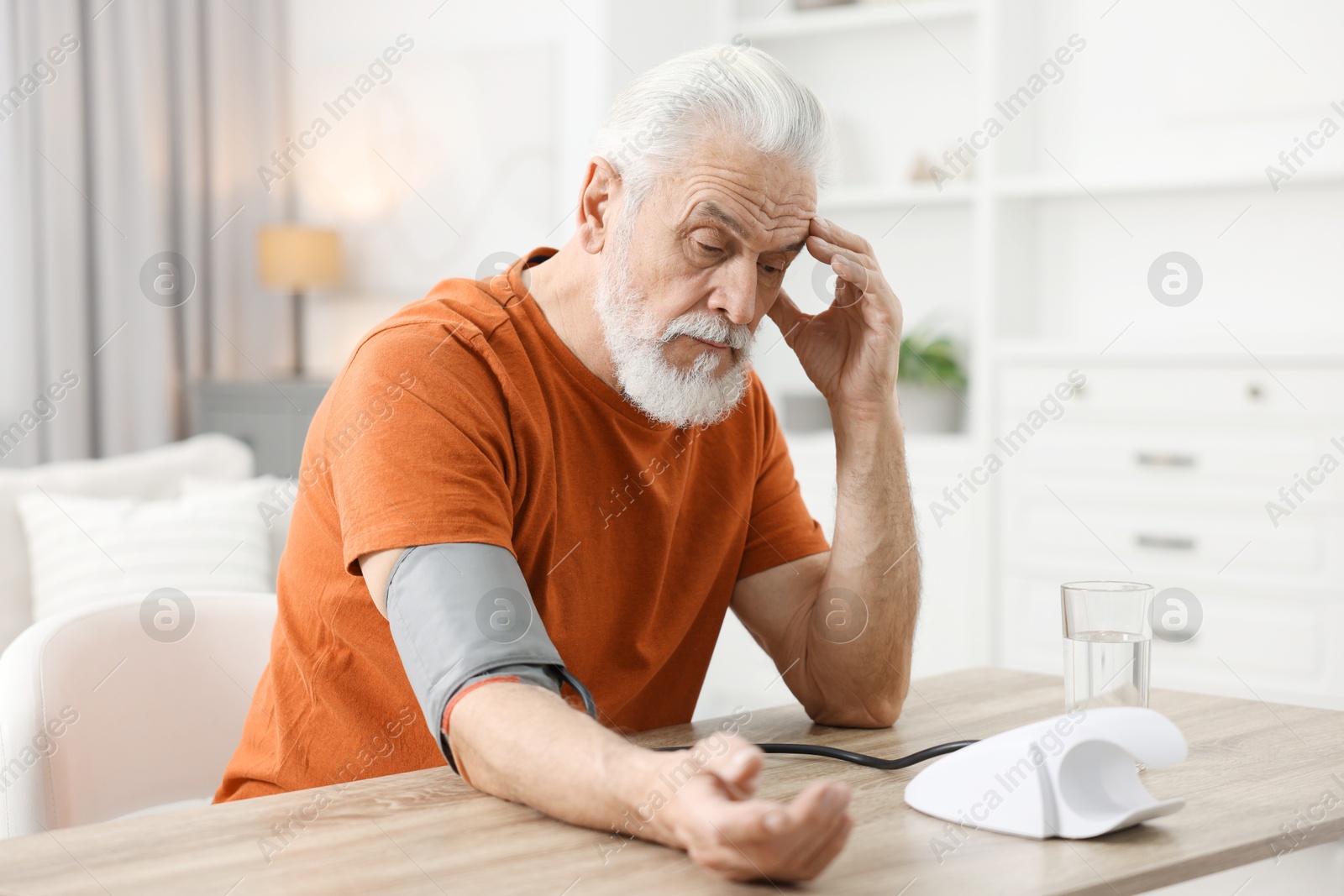 Photo of Worried senior man measuring blood pressure at table indoors