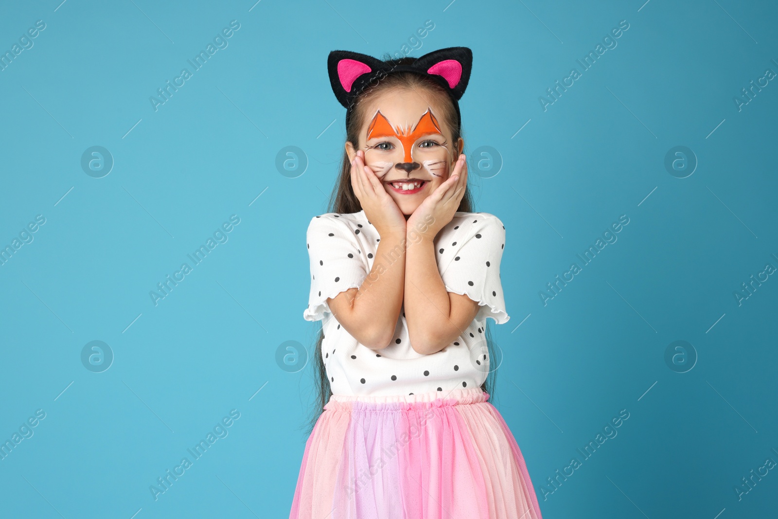 Photo of Cute girl with painted face and ears as cat on light blue background