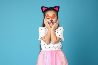 Photo of Cute girl with painted face and ears as cat on light blue background