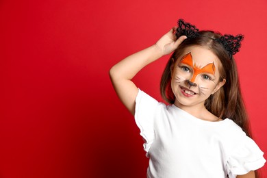 Photo of Cute girl with painted face and ears as cat on red background, space for text