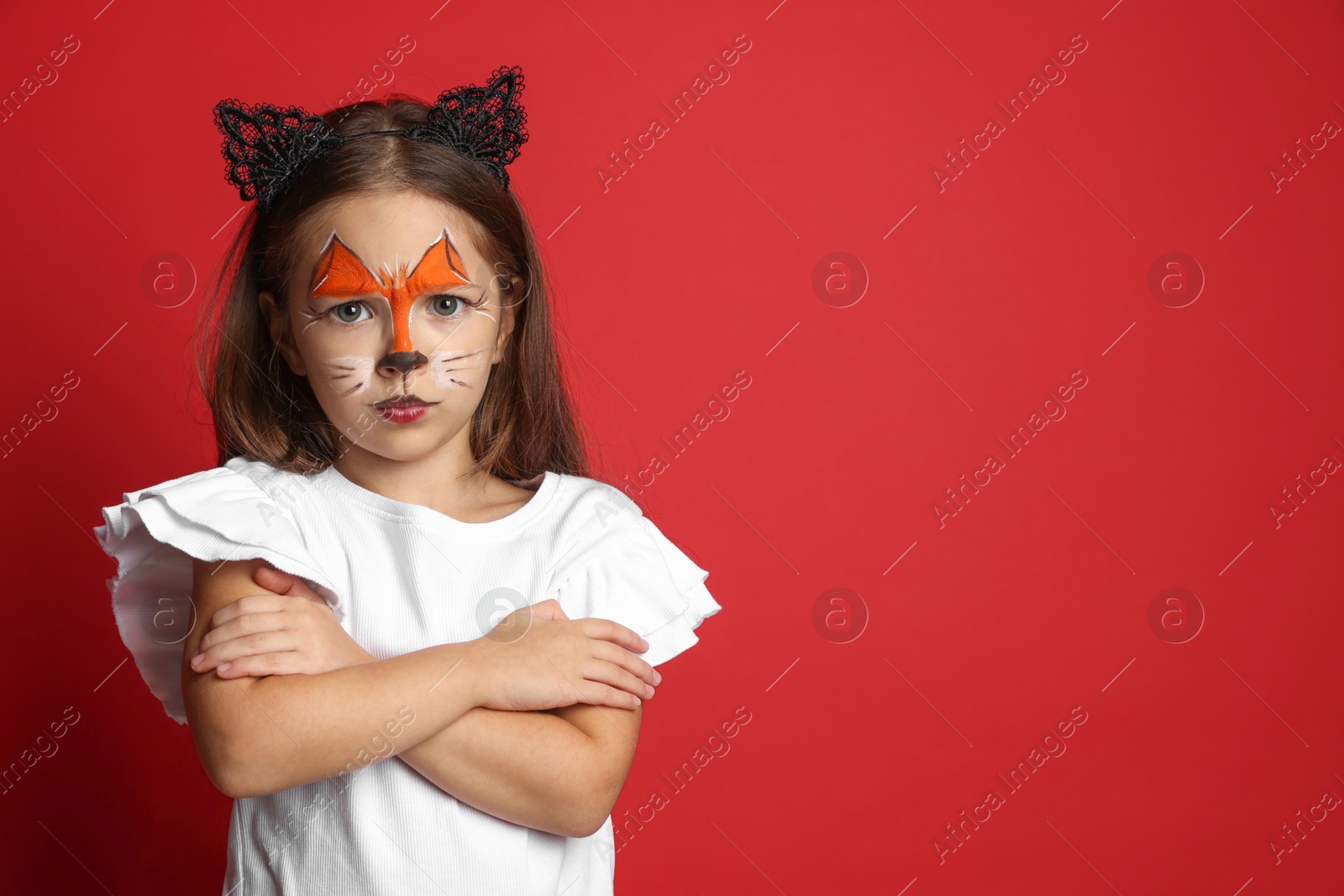 Photo of Cute girl with painted face and ears as cat on red background, space for text