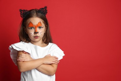 Photo of Cute girl with painted face and ears as cat on red background, space for text