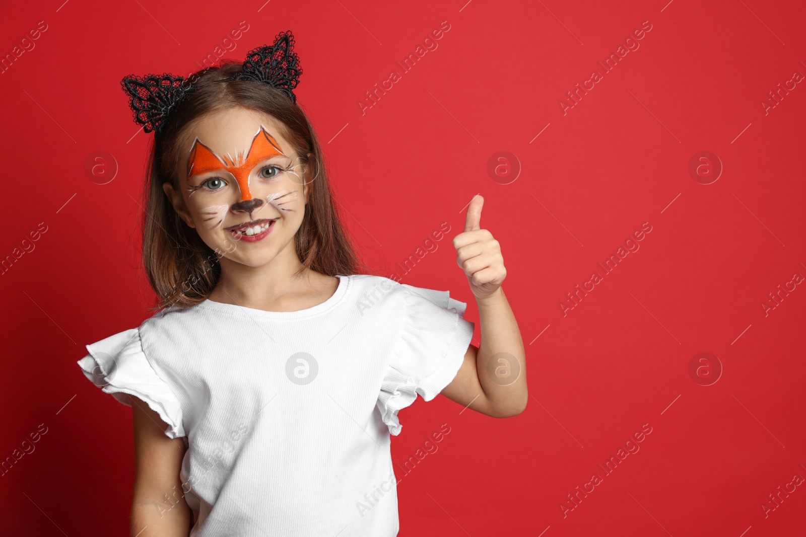 Photo of Cute girl with painted face and ears as cat showing thumbs up on red background, space for text