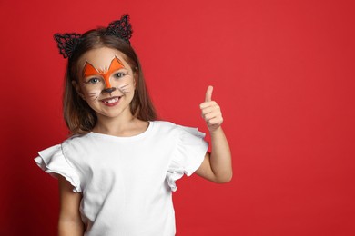 Photo of Cute girl with painted face and ears as cat showing thumbs up on red background, space for text