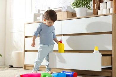 Photo of Little boy near cabinet with bottles of detergents at home. Child in danger