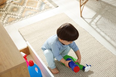 Photo of Little boy playing with bottles of detergents near cabinet at home. Child in danger