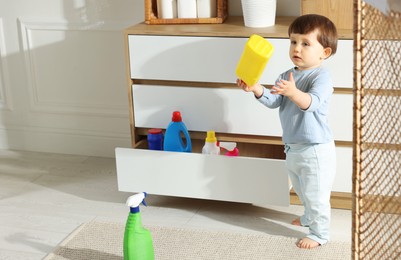 Photo of Little boy playing with bottle of detergent near cabinet at home, space for text. Child in danger