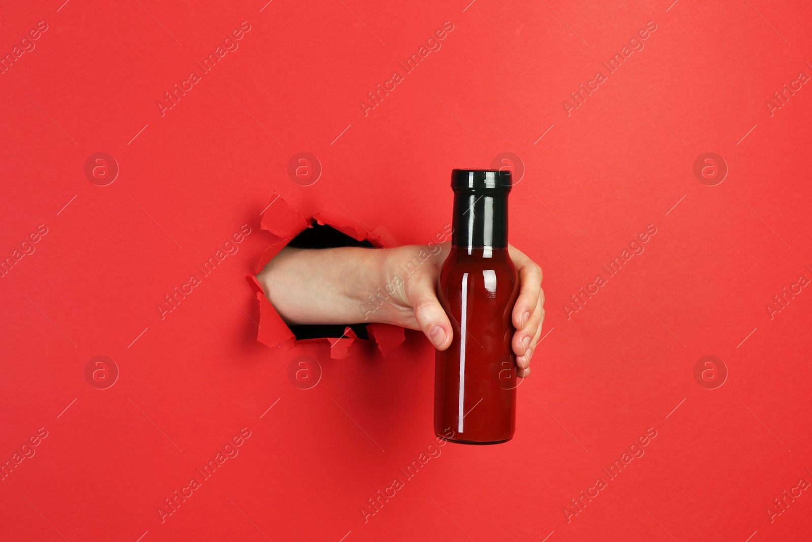 Photo of Woman holding bottle of ketchup through hole in torn red paper, closeup