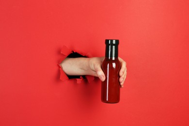 Photo of Woman holding bottle of ketchup through hole in torn red paper, closeup