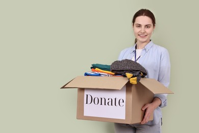 Photo of Woman holding donation box with clothes on pale olive background, space for text