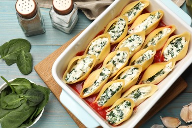 Photo of Delicious conchiglie pasta with ricotta cheese, spinach and tomato sauce in baking dish on light blue wooden table, flat lay