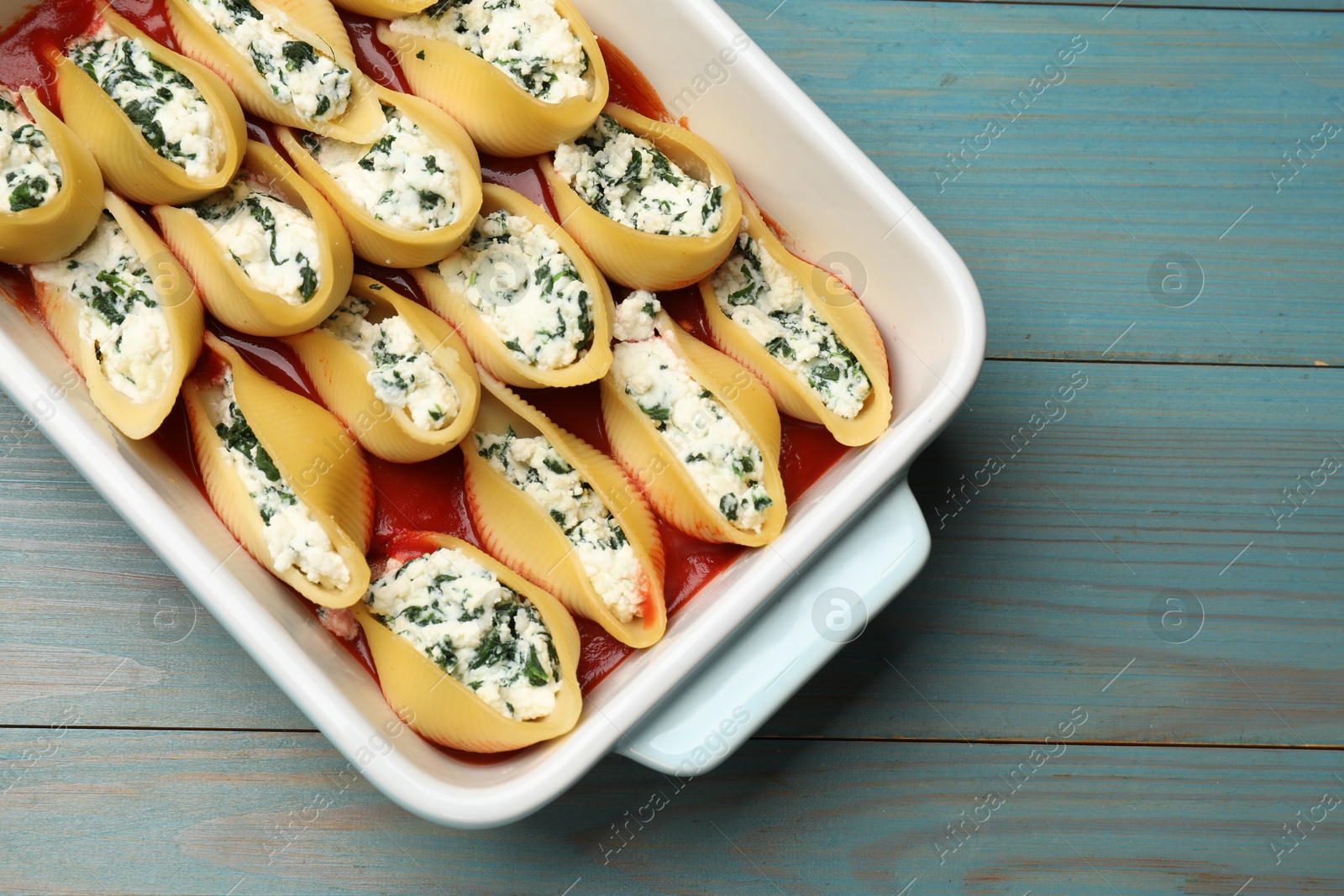 Photo of Delicious pasta with ricotta cheese, spinach and tomato sauce in baking dish on light blue wooden table, top view. Space for text