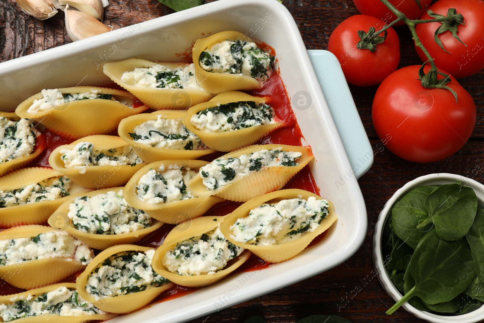 Photo of Delicious conchiglie pasta with ricotta cheese, spinach and tomato sauce in baking dish on wooden table, flat lay