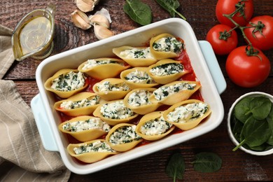 Photo of Delicious conchiglie pasta with ricotta cheese, spinach and tomato sauce in baking dish on wooden table, flat lay