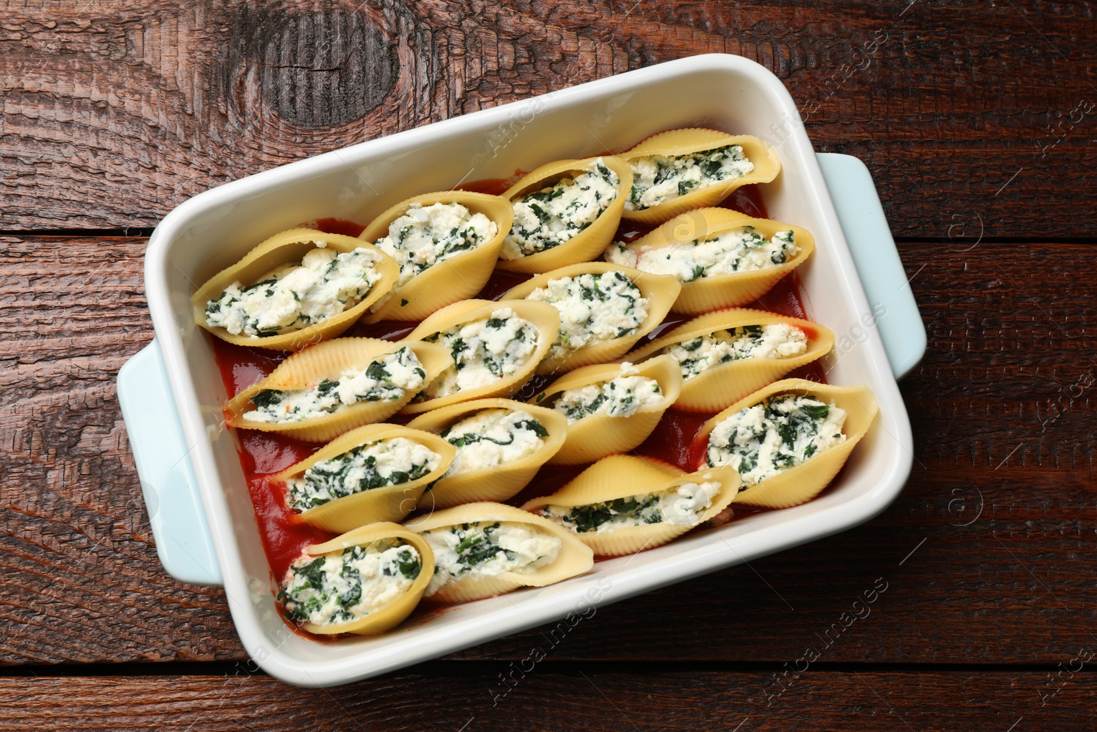 Photo of Delicious conchiglie pasta with ricotta cheese, spinach and tomato sauce in baking dish on wooden table, top view