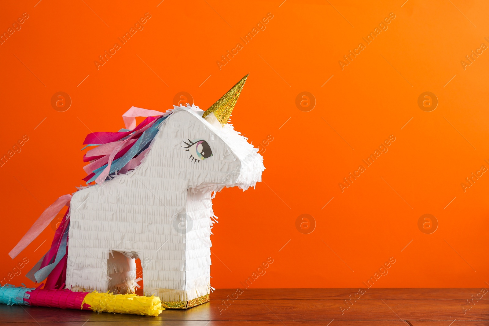 Photo of Bright pinata in shape of unicorn and stick on table against orange background. Space for text