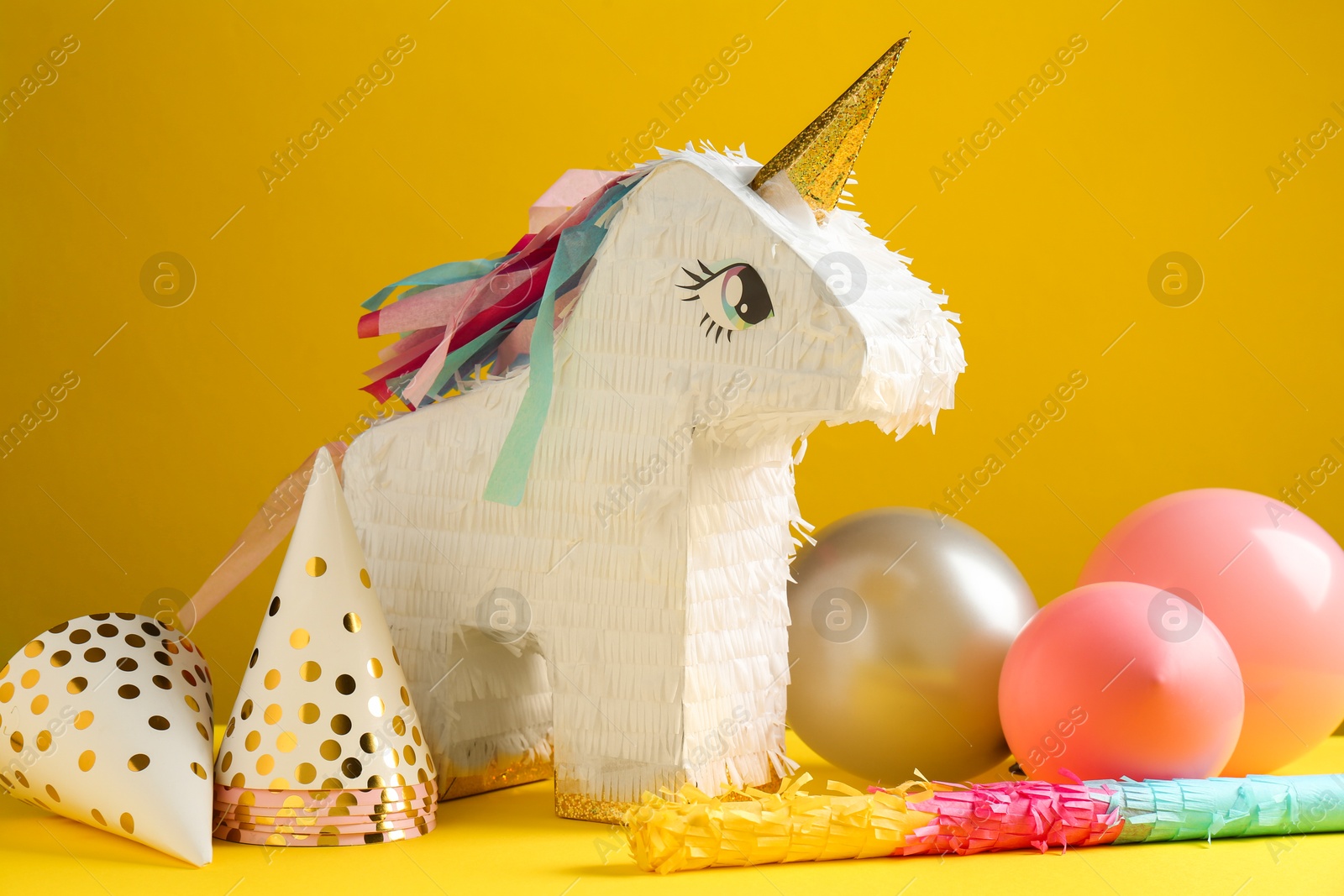 Photo of Bright pinata in shape of unicorn, stick, party hats and balloons on yellow background