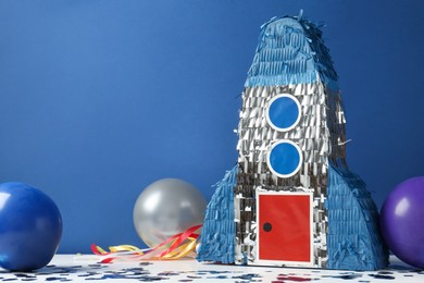 Photo of Bright pinata in shape of spaceship, balloons and confetti on table against blue background. Space for text