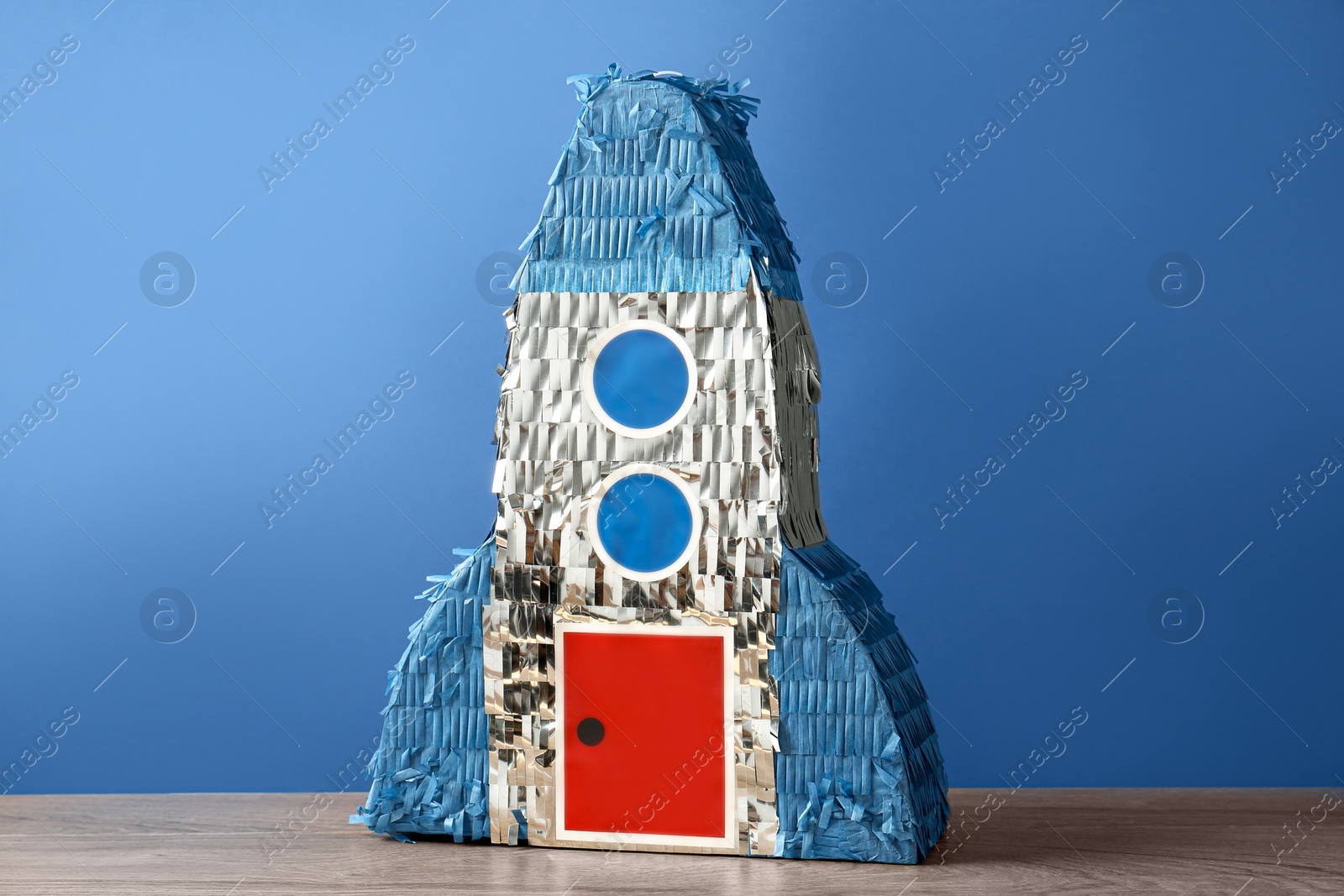 Photo of Beautiful pinata in shape of rocket on wooden table against blue background