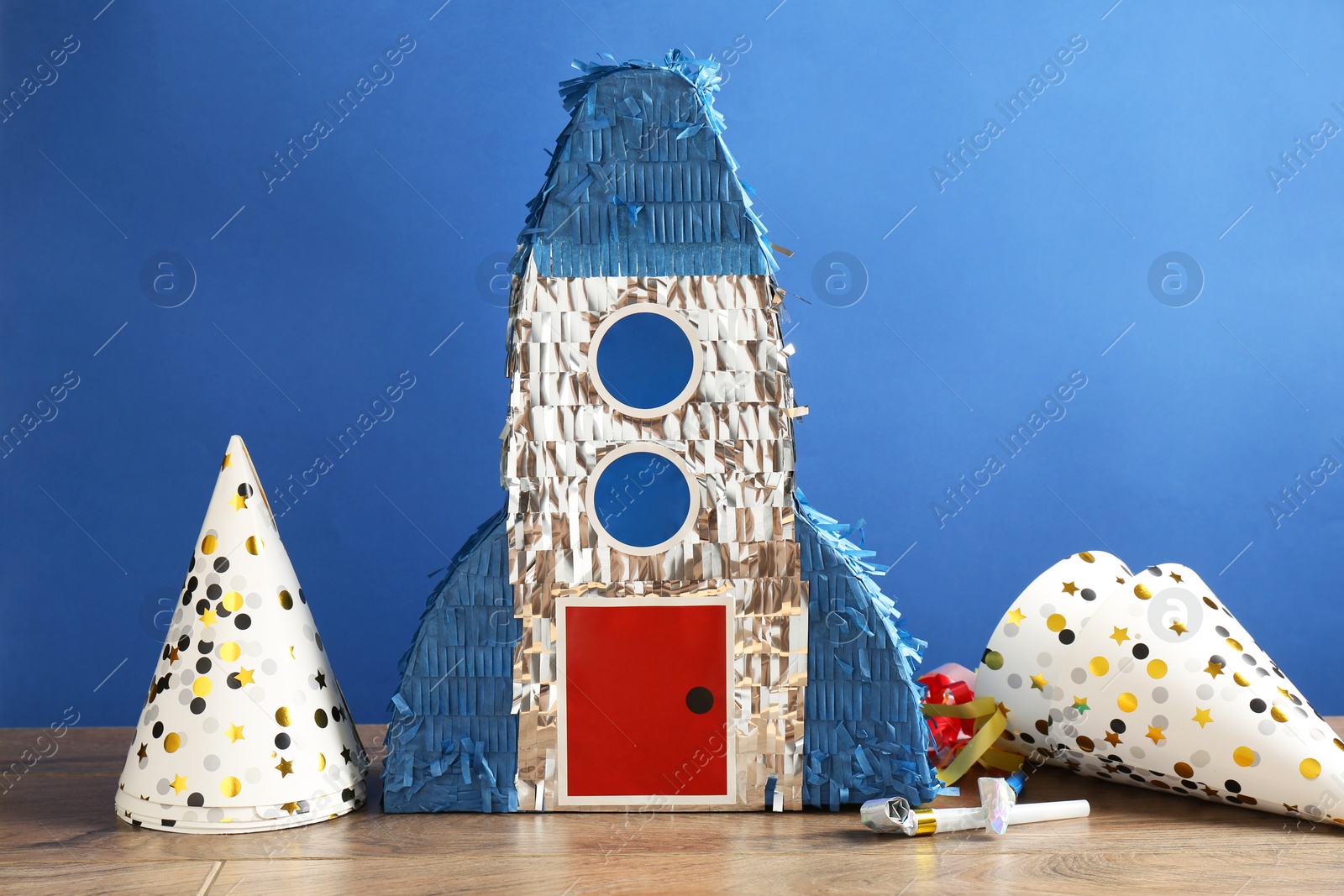 Photo of Beautiful pinata in shape of rocket, party cones and blowers on wooden table against blue background