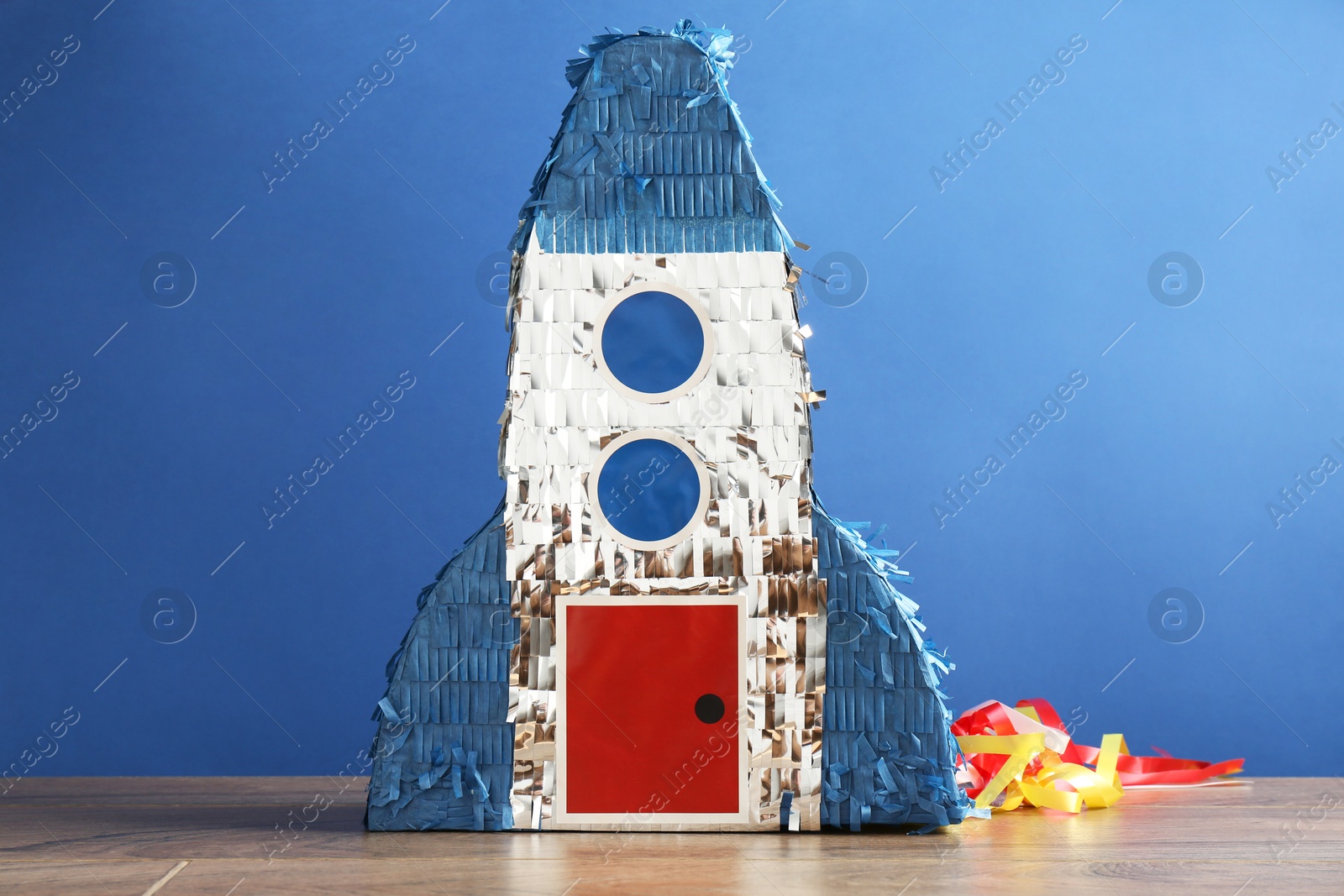 Photo of Beautiful pinata in shape of rocket on wooden table against blue background