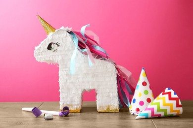 Photo of Beautiful pinata in shape of unicorn, party cones and blowers on wooden table against pink background