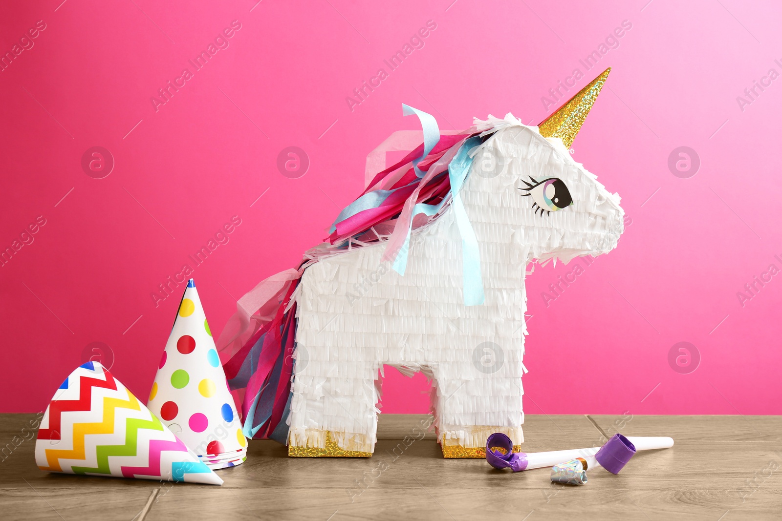 Photo of Beautiful pinata in shape of unicorn, party cones and blowers on wooden table against pink background