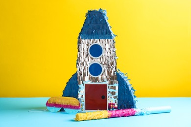 Photo of Beautiful pinata in shape of rocket, blindfold and stick on blue table against yellow background