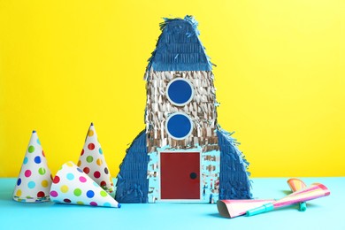 Photo of Beautiful pinata in shape of rocket, party cones and poppers on blue table against yellow background