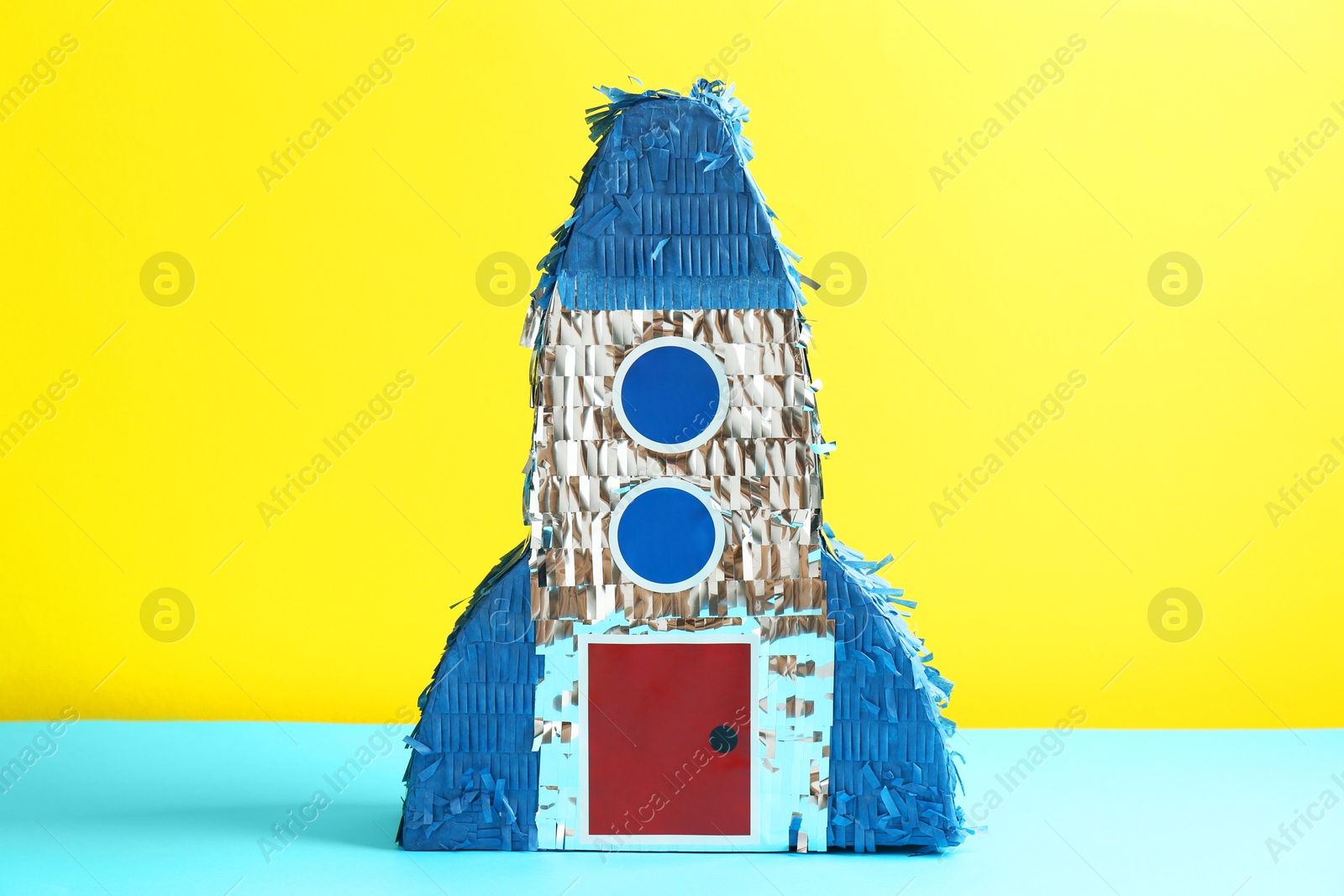 Photo of Beautiful pinata in shape of rocket on blue table against yellow background