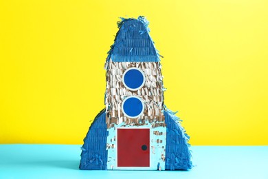 Photo of Beautiful pinata in shape of rocket on blue table against yellow background