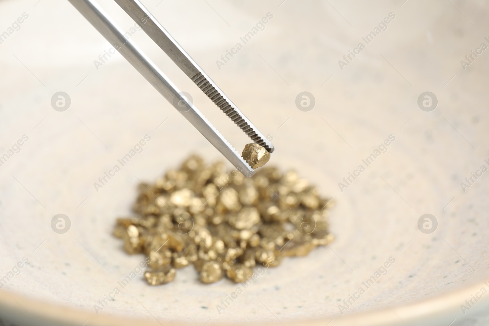 Photo of Tweezers with gold nugget above pile against blurred background, closeup