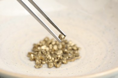 Photo of Tweezers with gold nugget above pile against blurred background, closeup