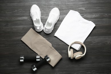 Photo of Stylish sportswear, white sneakers, dumbbells and headphones on wooden background, flat lay