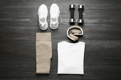 Photo of Stylish sportswear, white sneakers, dumbbells and headphones on wooden background, flat lay