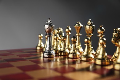 Photo of Silver king in front of golden chess pieces on chessboard against grey background, closeup. Competition concept