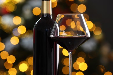 Photo of Red wine in glass and bottle against blurred Christmas lights, closeup