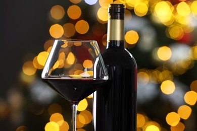 Photo of Red wine in glass and bottle against blurred Christmas lights, closeup