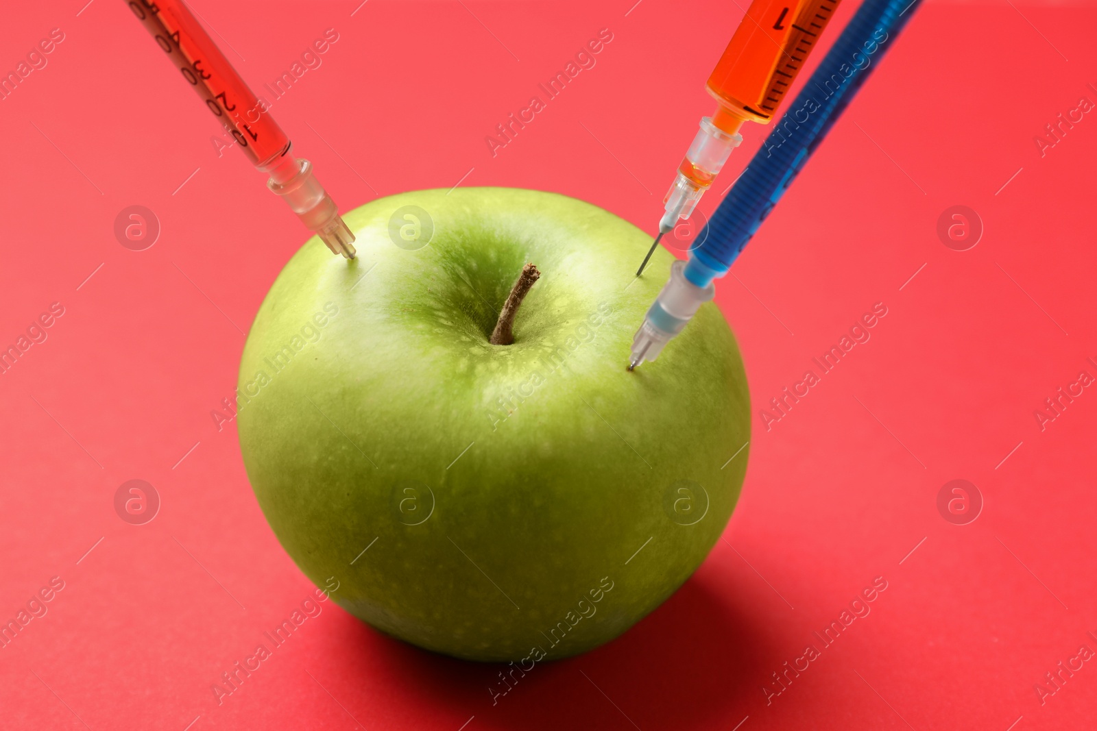 Photo of GMO concept. Green apple with different syringes on red background, closeup