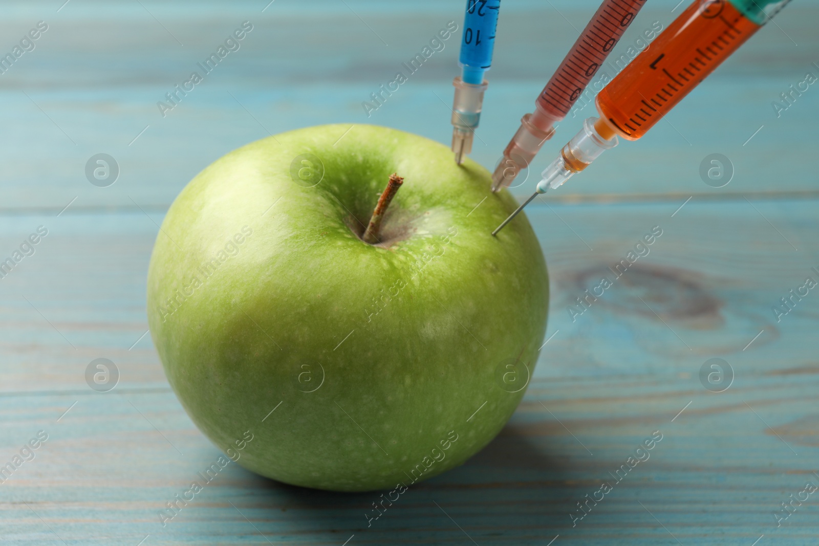 Photo of GMO concept. Green apple with syringes on light blue wooden table, closeup