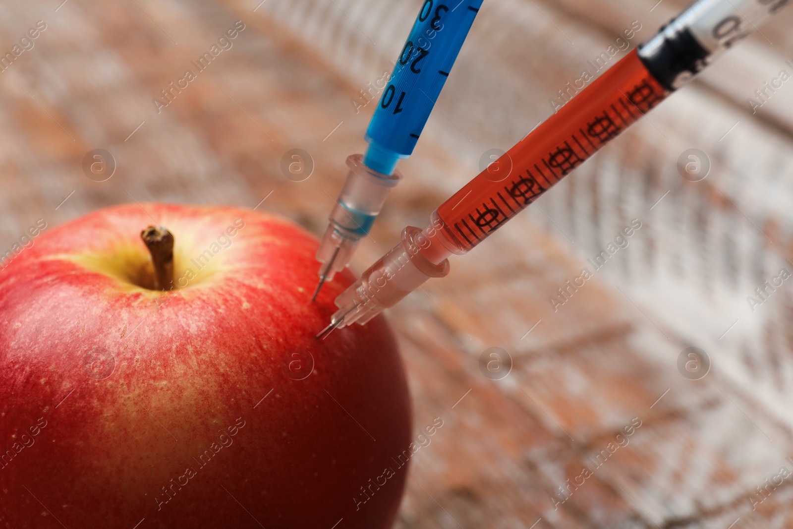 Photo of GMO concept. Red apple with syringes on blurred background, closeup