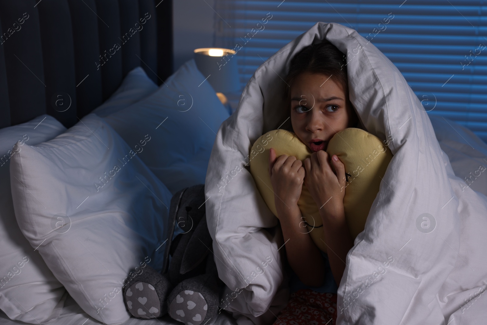 Photo of Afraid girl with pillow under duvet on bed at night