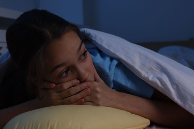 Photo of Afraid girl with pillow under duvet on bed at night