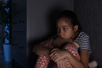 Photo of Scared girl with teddy bear hiding behind chest of drawers in her room at night