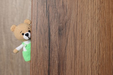 Photo of Cute knitted bear peeking out of wooden door, space for text